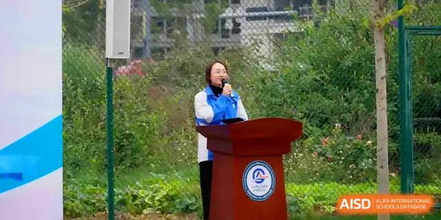 A speaker addresses the audience from a wooden podium during an outdoor ceremony at The Guandu Experimental School Affiliated To Shanghai Normal University.