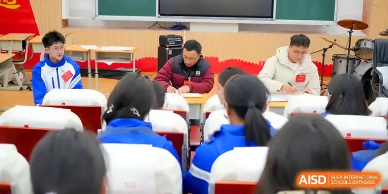 A teacher in a red jacket takes notes while sitting at a desk among students in a classroom at The Middle School Affiliated to Sichuan University.