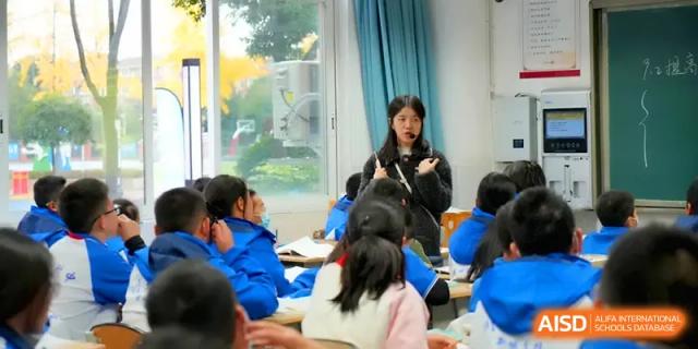 A female instructor in a dark coat stands among students in a bright classroom while leading an educational discussion at The Middle School Affiliated to Sichuan University.