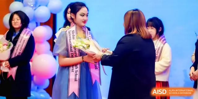 A student in a formal dress receives a celebratory bouquet of flowers from an official on stage during an awards ceremony at The Middle School Affiliated to Sichuan University.