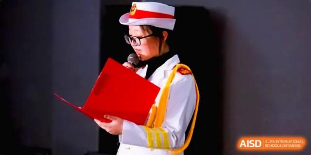 A student in a white uniform and red sash reads from a red folder during a formal school presentation on stage at The Middle School Affiliated to Sichuan University.