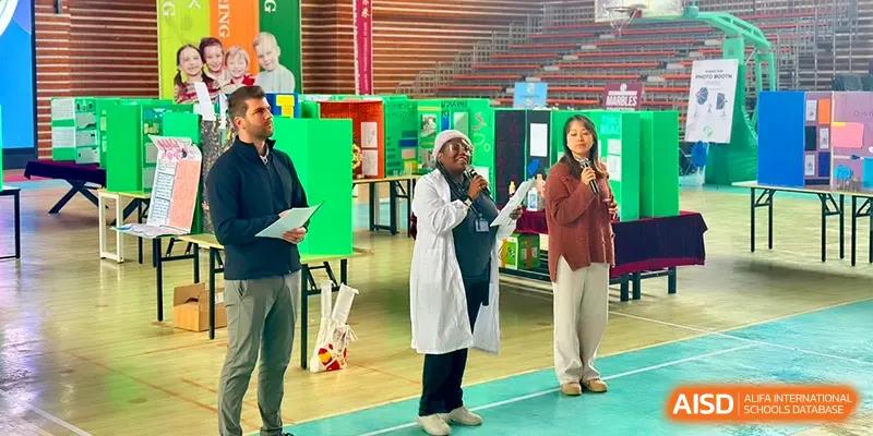 Three presenters speak into microphones in front of science project boards at Xiwai International School.