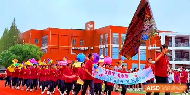 A large procession of students in red shirts marches past a bright red school building during a festival at Xiwai International School.