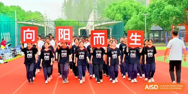 Students in black athletic uniforms march in formation while holding large red square signs at Xiwai International School.