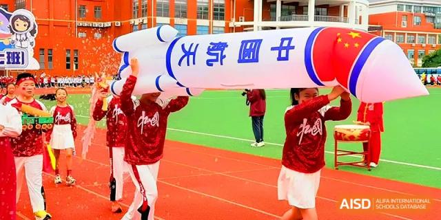 Students work together to carry a large inflatable rocket model across a green sports field at Xiwai International School.