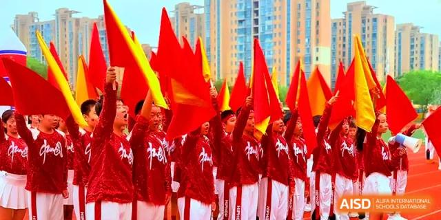 A group of students in red and white uniforms marches while carrying large red flags during a ceremony at Xiwai International School.