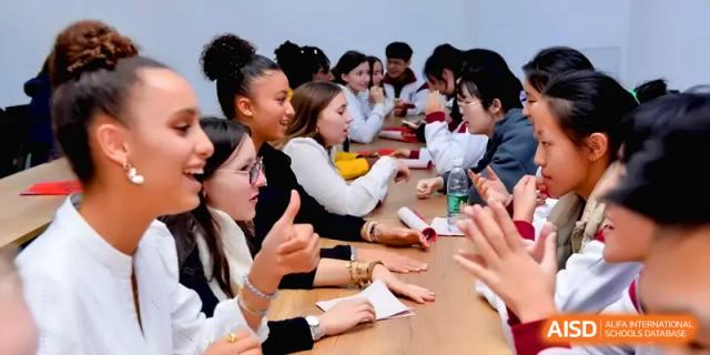 A diverse group of students sits around a long wooden table, talking and gesturing during a group session at Xuzhou No.1 Middle School.