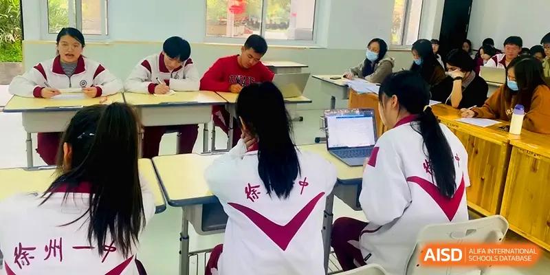 A student in a white and maroon jacket works on a laptop at a desk in a well-lit classroom at Xuzhou No.1 Middle School.