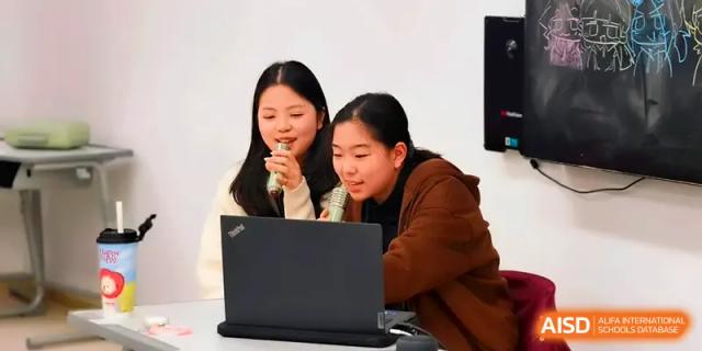 Two girls smile while looking at a laptop and singing into a microphone during a festive event at Xuzhou No.1 Middle School.