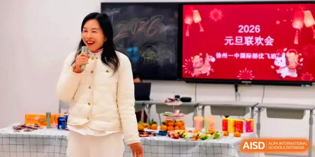 A teacher in a white jacket smiles during a New Year party featuring festive snacks and digital banners at Xuzhou No.1 Middle School.