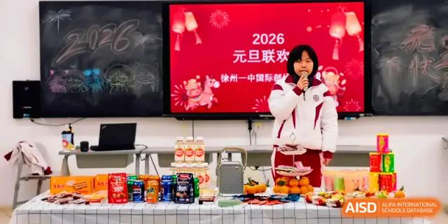 A student speaks into a microphone next to a table of snacks and a 2026 New Year digital display at Xuzhou No.1 Middle School.