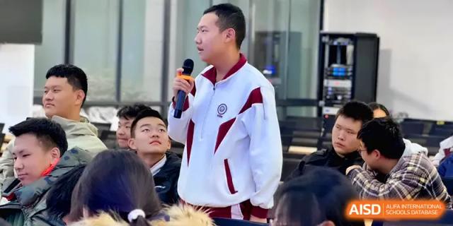 A student in a white and maroon tracksuit stands and speaks into a microphone during a group session at Xuzhou No.1 Middle School.