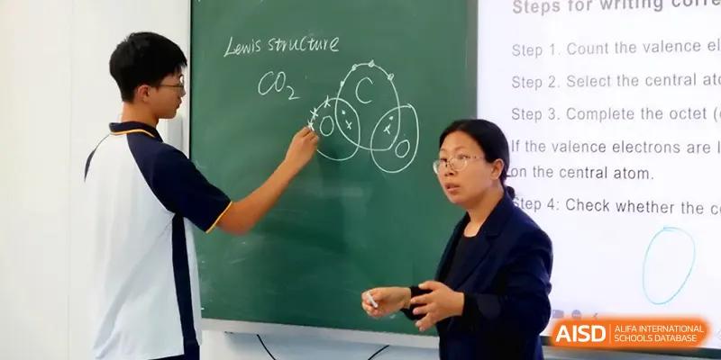 A teacher explains the Lewis structure of CO2 on a chalkboard while a student points to the diagram during a science lesson at Beijing Xinxuedao Linchuan School.