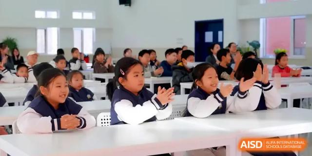 Students sit at long white tables and clap their hands during a fun and engaging classroom activity at Beijing Xinxuedao Linchuan School.
