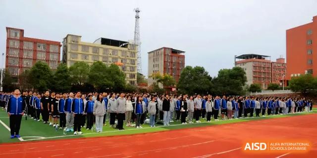 A large group of students in uniform stands in formation on the red athletic track for an outdoor assembly at Changsha Mingda Middle School.