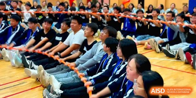 A line of students sits on the ground, holding a long orange pole during a fun tug-of-war style game at Changsha Mingda Middle School.