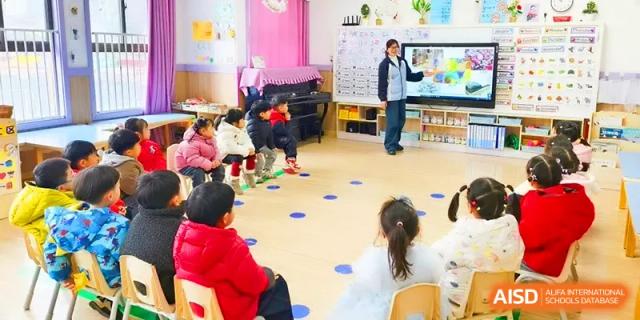 A teacher in a white coat stands before a digital screen in a brightly colored preschool classroom during a morning lesson at Walton-Hefei School.