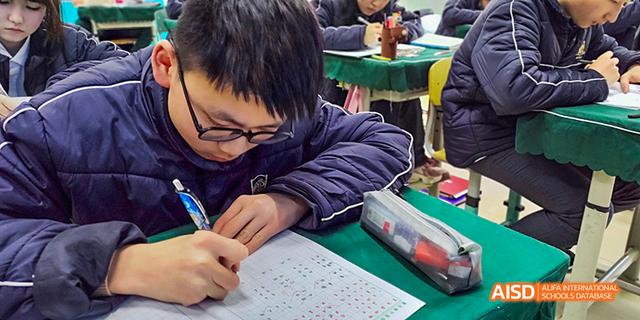 A student wearing glasses concentrating on writing answers on a test or worksheet during a class at Maple Leaf International School Chongqing.
