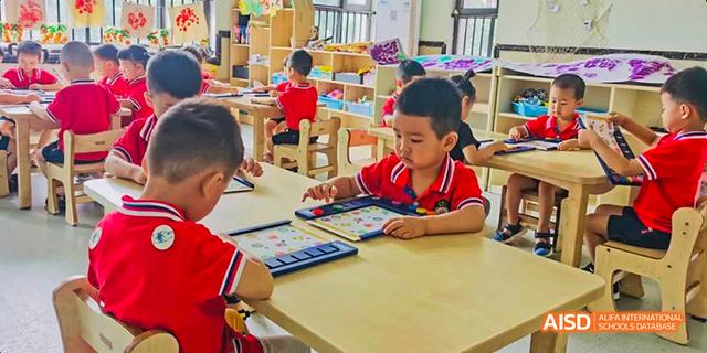 Young students in red shirts seated at low wooden desks and working with learning tools or tablets in an early childhood classroom at Maple Leaf International School Chongqing.
