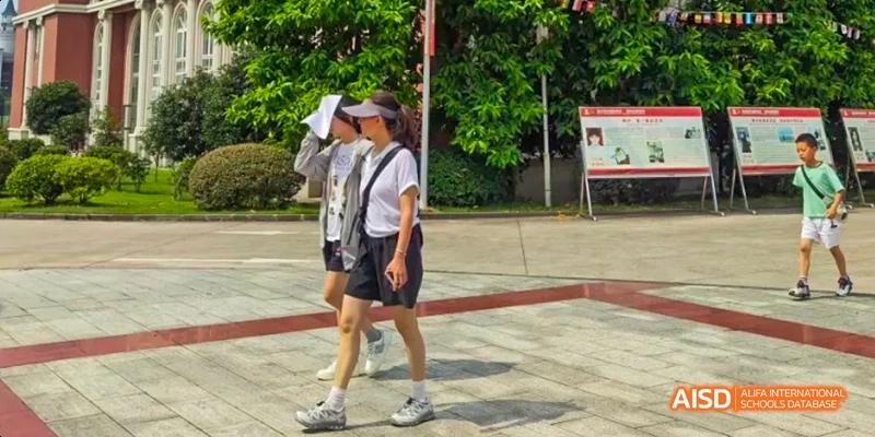 Two students walking on a paved area, one wearing a white shirt and black shorts with a visor, on the campus of Maple Leaf International School Chongqing.