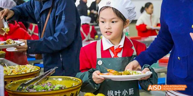 A young female student wearing a chef's hat and apron serving food on a plate, possibly at a cooking event, at Maple Leaf International School Chongqing.