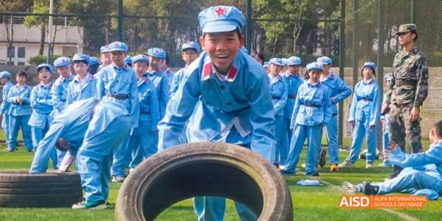 A smiling male student in a blue military-style uniform pulling or pushing a large black tire during an outdoor training exercise at Maple Leaf International School Chongqing.