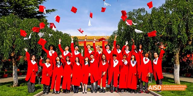 Canadian International School of Beijing graduates in red robes throwing their caps into the air outdoors in front of a building.