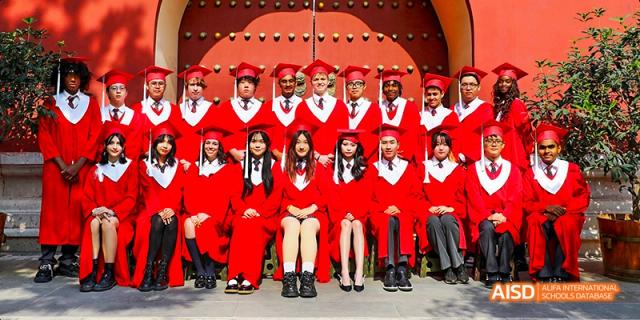 Canadian International School of Beijing graduates in red robes and caps seated in a front row for a group photo, with other graduates standing behind them.