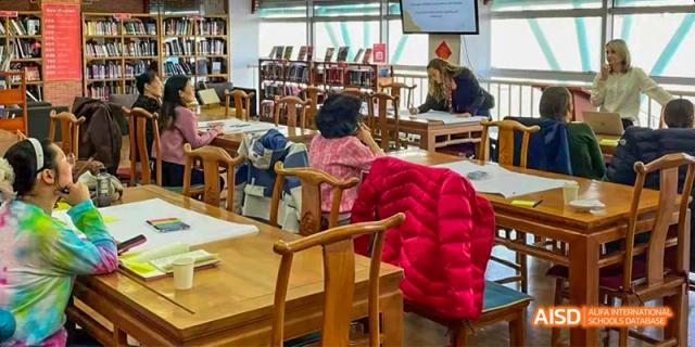 Canadian International School of Beijing students working or studying at individual wooden desks in a library or classroom filled with bookshelves.