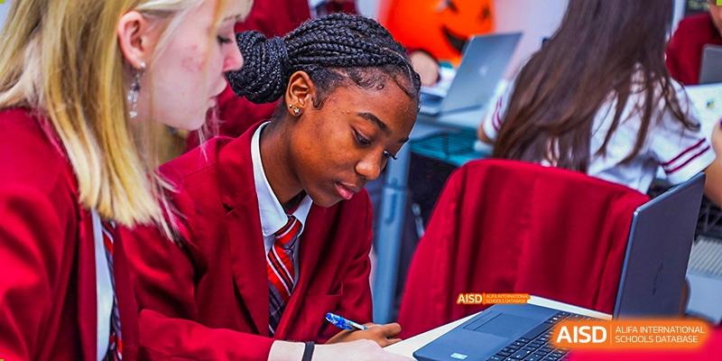 Canadian International School of Beijing female student with braided hair, wearing a red uniform, writing in a notebook with a laptop in front of her.