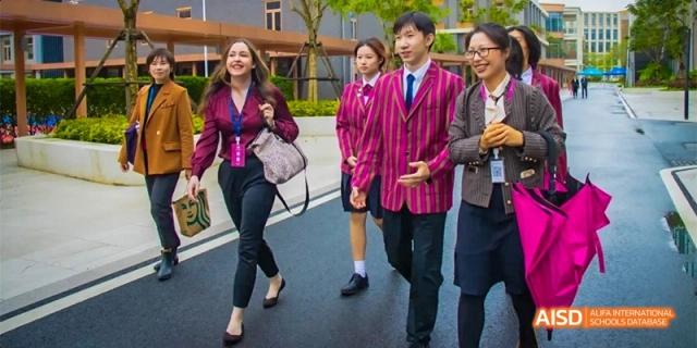 A student at Fettes College Guangzhou, wearing a distinctive pink and maroon striped blazer, walking outdoors with other staff or students.