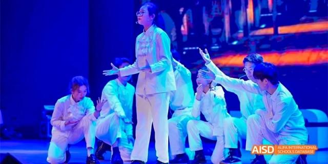 A performer on stage at Fettes College Guangzhou, dressed in white traditional attire, singing or acting during a cultural performance with a group.