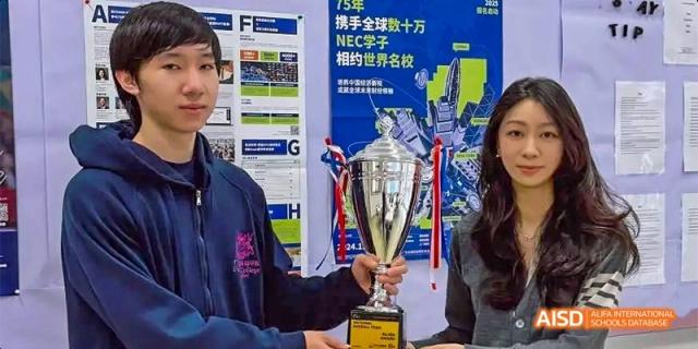 A student from Fettes College Guangzhou proudly holding a large silver trophy at a school event, with academic posters visible in the background.