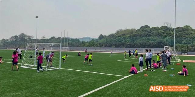 Students from Fettes College Guangzhou playing a game of soccer on a wide, green turf field with hills and trees visible in the distance.