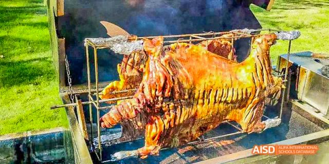 A large piece of whole roasted meat, possibly a pig or lamb, cooking over an open fire at a culinary event at Fettes College Guangzhou.