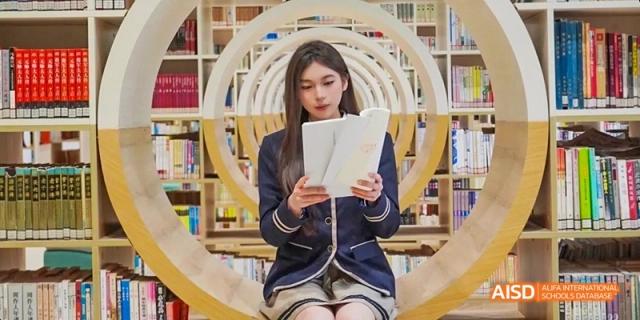 A Guangzhou Huamei International School student in uniform sitting and reading a book within a large, brightly lit library or study space.