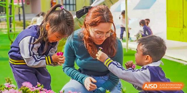 A teacher or staff member at Guangzhou Huamei International School kneeling on the grass, smiling, and holding a microphone up to a small child.