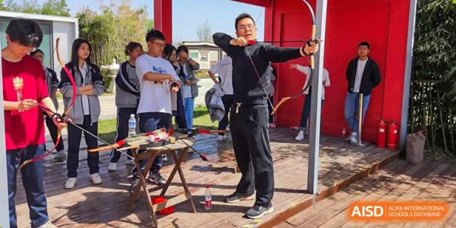 A male teacher at Zhengzhou No.9 High School International Department practices archery with a bow and arrow outdoors, with students watching nearby.