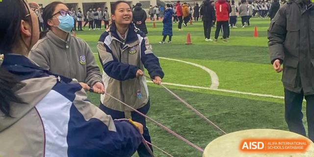 A female student at Zhengzhou No.9 High School International Department smiles while pulling a rope during an outdoor sports day or team competition on a grass field.