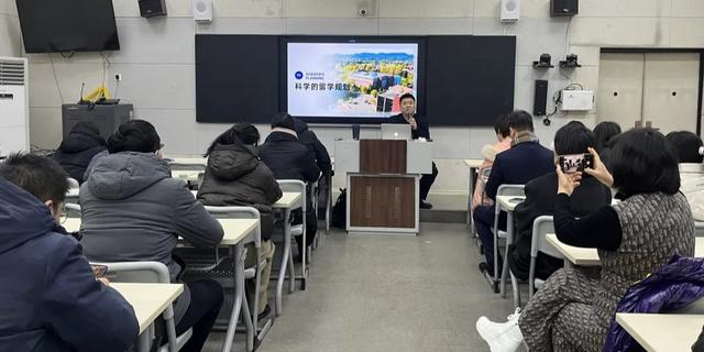 A teacher instructing a class in a lecture hall setting with a large projection screen at Zhengzhou No.9 High School International Department.