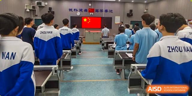Students from Zhounan High School sitting in an auditorium or large classroom, facing a stage with a red banner and screens.