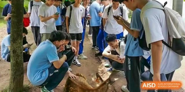 Students from Zhounan High School gathered outdoors, with some kneeling to interact with a small animal, possibly a goat or sheep.
