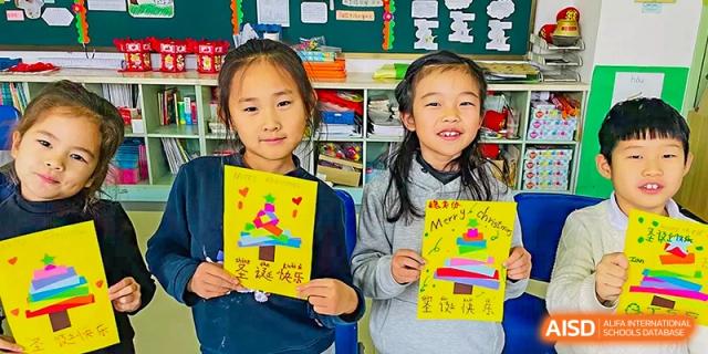 Two young female students smile and hold up drawings or art projects in a classroom at Qingdao No.1 International School of Shandong Province (QISS).