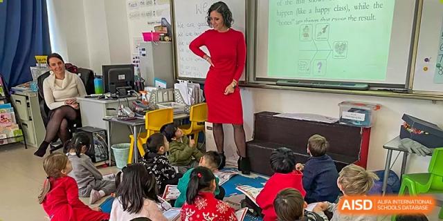A teacher in a red dress stands and talks to a group of students sitting on the floor in a classroom at Qingdao No.1 International School of Shandong Province (QISS).