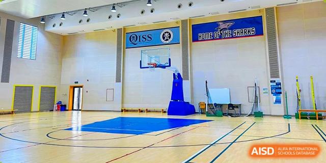 A blue basketball court with a hoop and a banner on the wall in a gymnasium at Qingdao No.1 International School of Shandong Province (QISS).