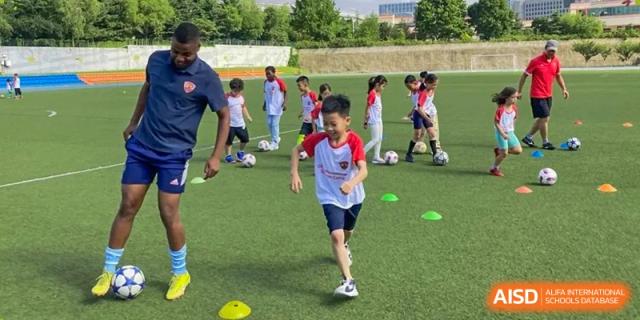 A student in a red and white uniform runs with a soccer ball on a green field at Qingdao No.1 International School of Shandong Province (QISS).