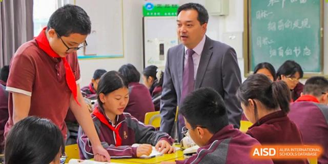 A male teacher in a suit assisting a student with their work in a busy classroom setting at Yichang Tianwen International School.