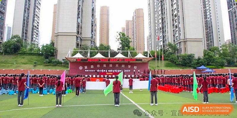 A wide view of a school event set up on a sports field with banners, students, and apartment buildings in the background at Yichang Tianwen International School.