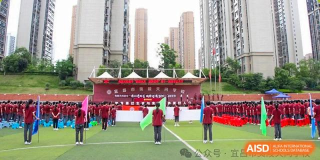 A wide view of a school event set up on a sports field with banners, students, and apartment buildings in the background at Yichang Tianwen International School.