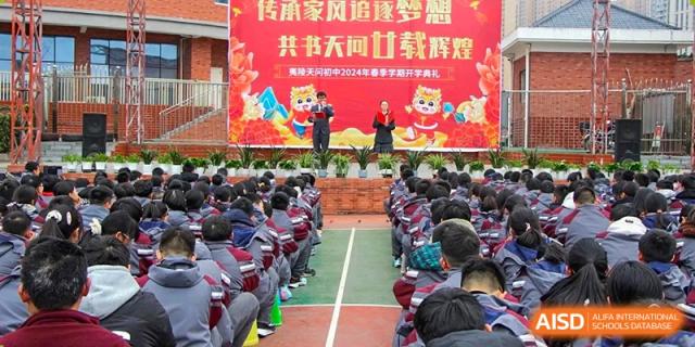 Students seated in formation outdoors, listening to a speaker on a stage during a school event or ceremony at Yichang Tianwen International School.